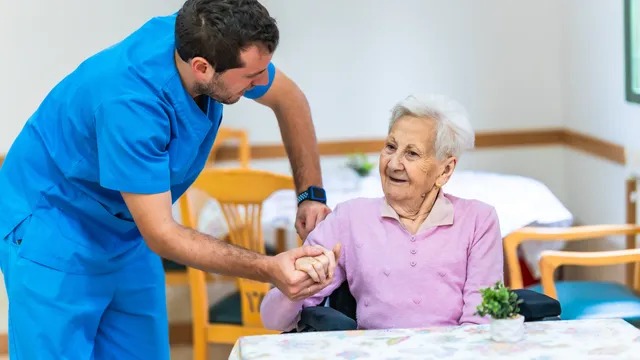 A care worker in uniform speaking with an older man seated in a wheelchair